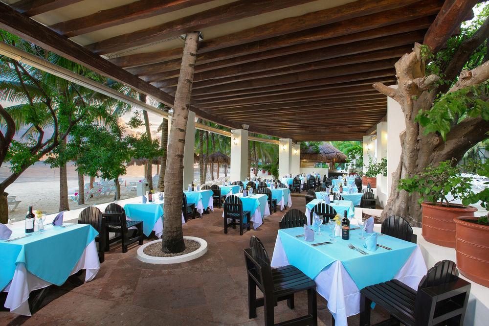 Grand Oasis Palm Ξενοδοχείο Κανκούν Εξωτερικό φωτογραφία The photo shows an outdoor dining area of a restaurant. The space features several tables covered with light blue tablecloths, each accompanied by black wooden chairs. The area is shaded by a wooden roof supported by columns, and there are leafy tree