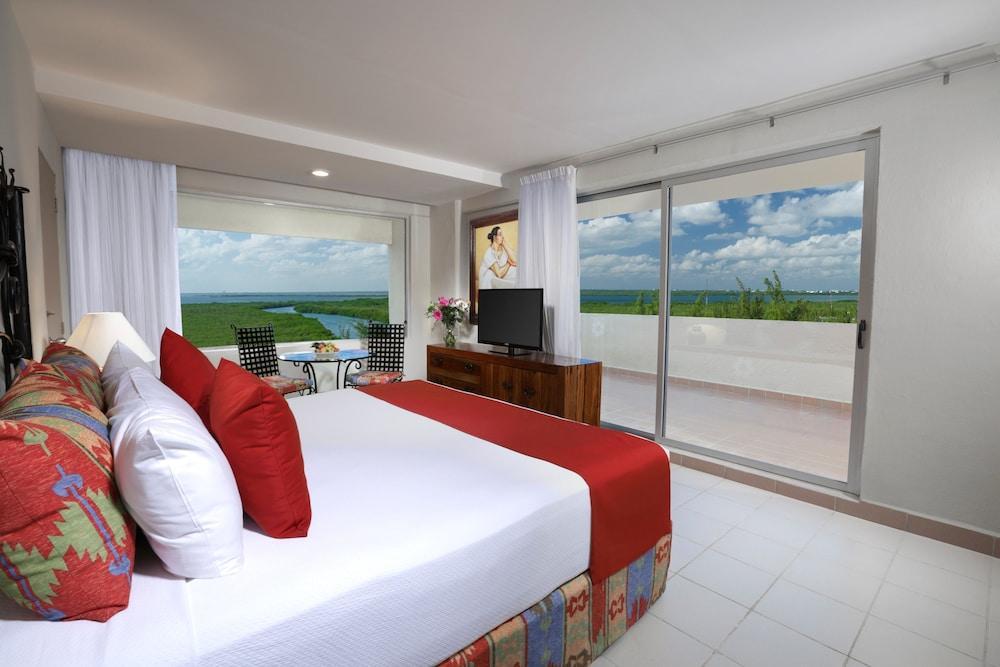 Grand Oasis Palm Ξενοδοχείο Κανκούν Εξωτερικό φωτογραφία The photo shows a modern hotel room featuring a large bed with white linens and red accents. There are decorative pillows on the bed, and a bedside table with a lamp. To the side, there’s a large window with white curtains that offers a view of a bal