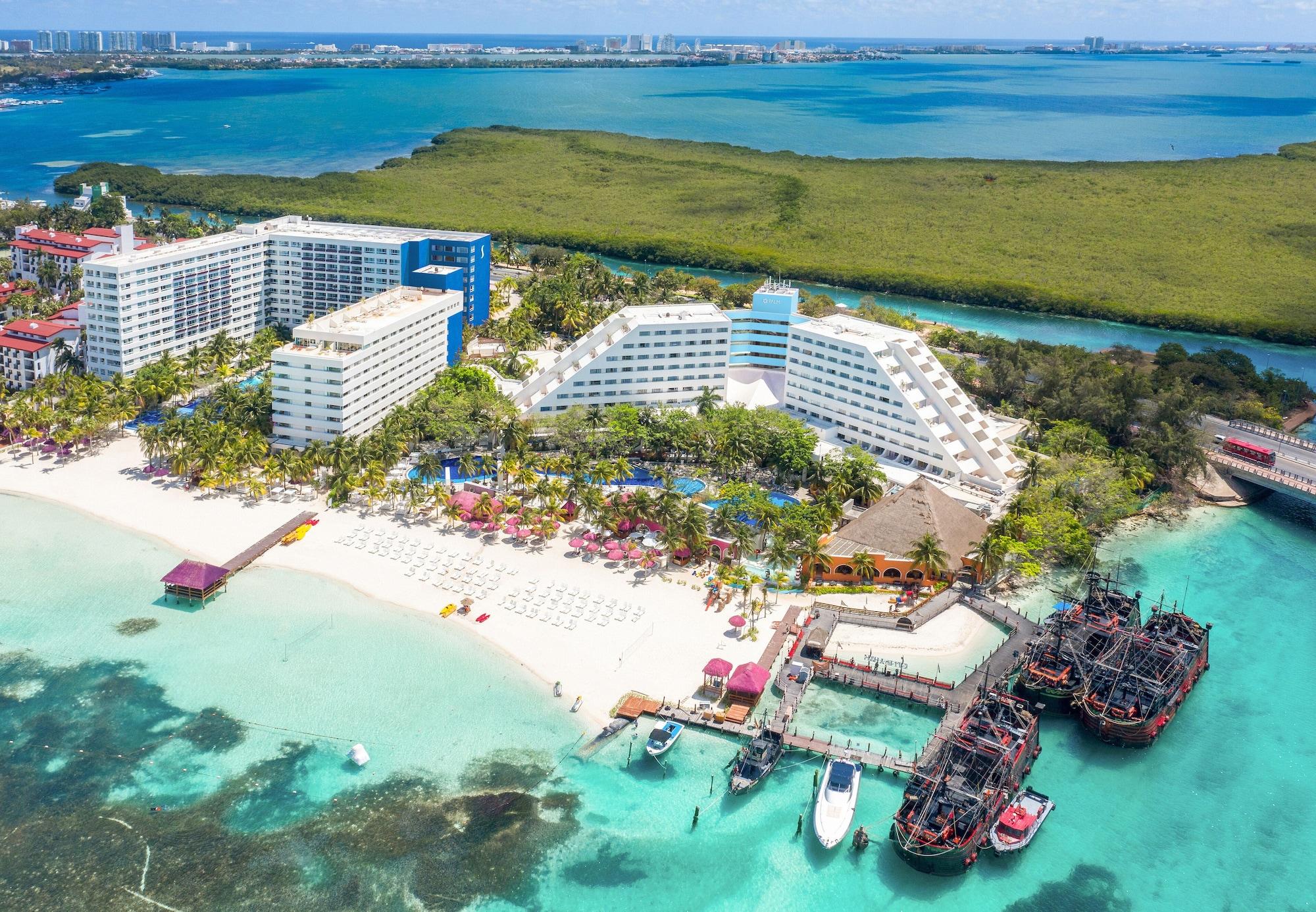 Grand Oasis Palm Ξενοδοχείο Κανκούν Εξωτερικό φωτογραφία The photo shows a beautiful coastal resort area. In the foreground, there is a sandy beach with lounge chairs and umbrellas, while the clear turquoise waters are visible. The resort features large white and blue buildings, with lush greenery surround