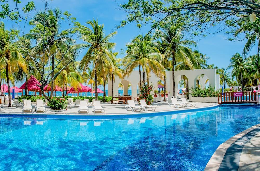 Grand Oasis Palm Ξενοδοχείο Κανκούν Εξωτερικό φωτογραφία The photo depicts a tropical resort scene featuring a large, inviting swimming pool surrounded by palm trees. The water in the pool is bright blue, reflecting the clear sky above. There are several white lounge chairs arranged around the pool area, p