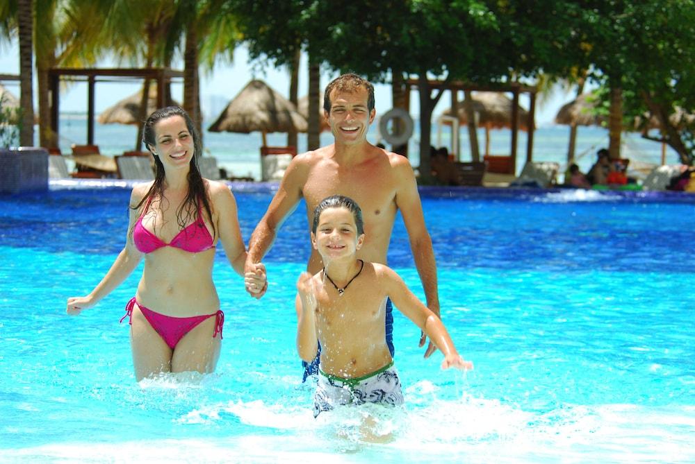 Grand Oasis Palm Ξενοδοχείο Κανκούν Εξωτερικό φωτογραφία The photo depicts a family enjoying a day at a pool. A woman in a pink bikini and a man are holding hands, while a boy runs happily in the water beside them. The pool is bright blue, and the background features palm trees and thatched huts, suggestin