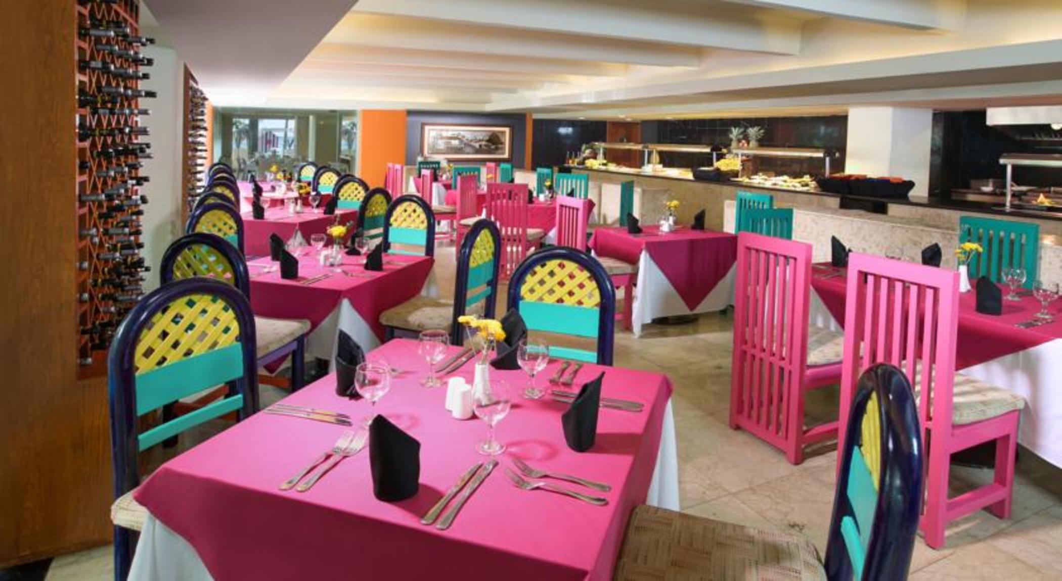 Grand Oasis Palm Ξενοδοχείο Κανκούν Εξωτερικό φωτογραφία The photo shows a vibrant restaurant interior featuring colorful decor. There are multiple tables covered with bright pink tablecloths. Each table is equipped with utensils, napkins, and a small centerpiece, possibly a flower. The chairs are painted 
